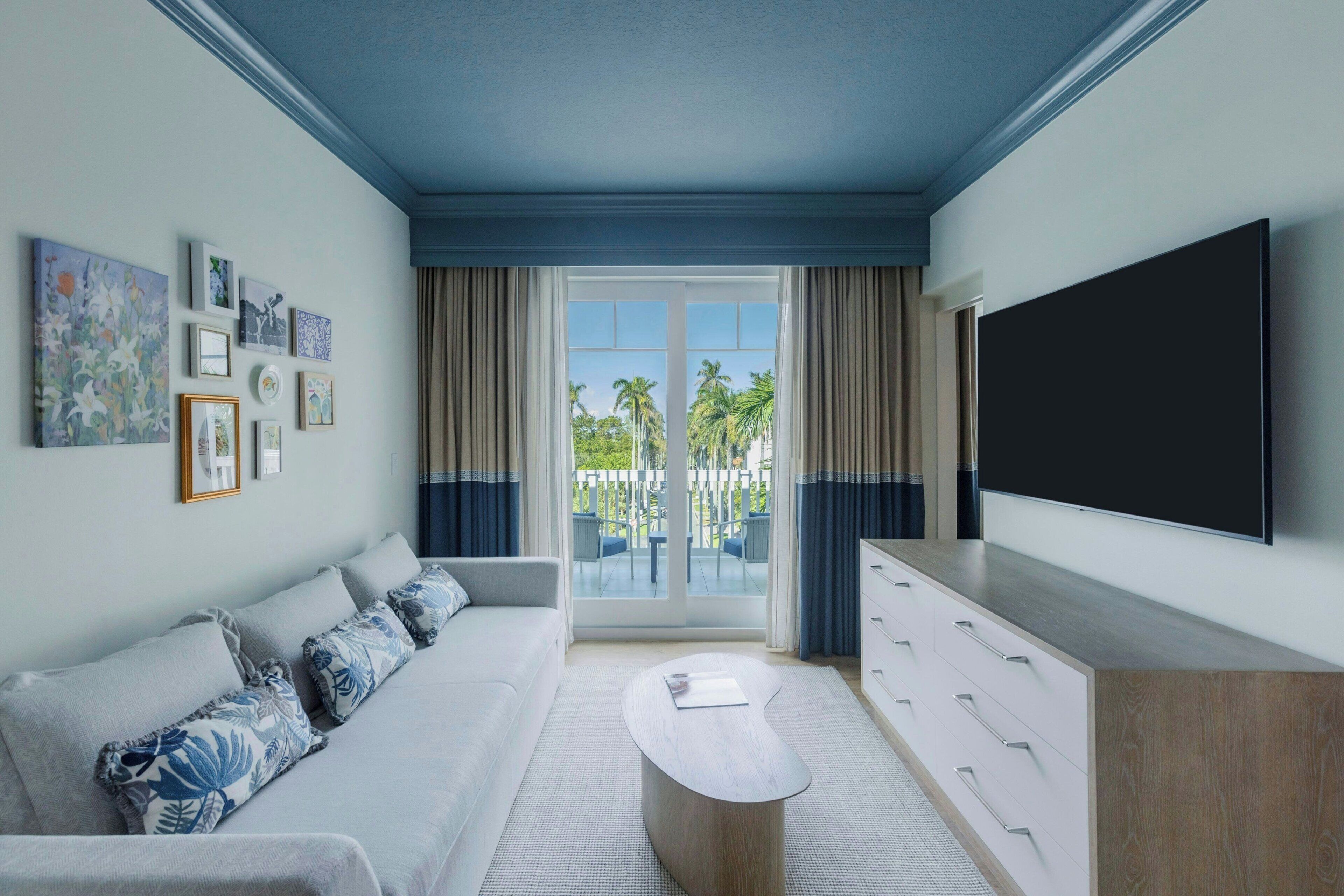 Een moderne hotelkamer met een grote flatscreen televisie, een bank met decoratieve kussens, een bijzettafeltje en uitzicht op palmbomen door de glazen deur.