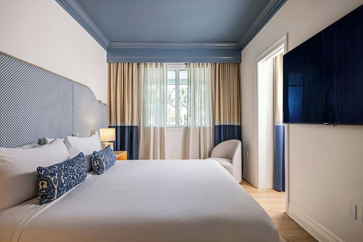 Ein Hotelzimmer mit einem großen Bett, einem Kopfteil, einem Sessel, einem Fenster mit Vorhängen und einem an der Wand befestigten Fernseher.
