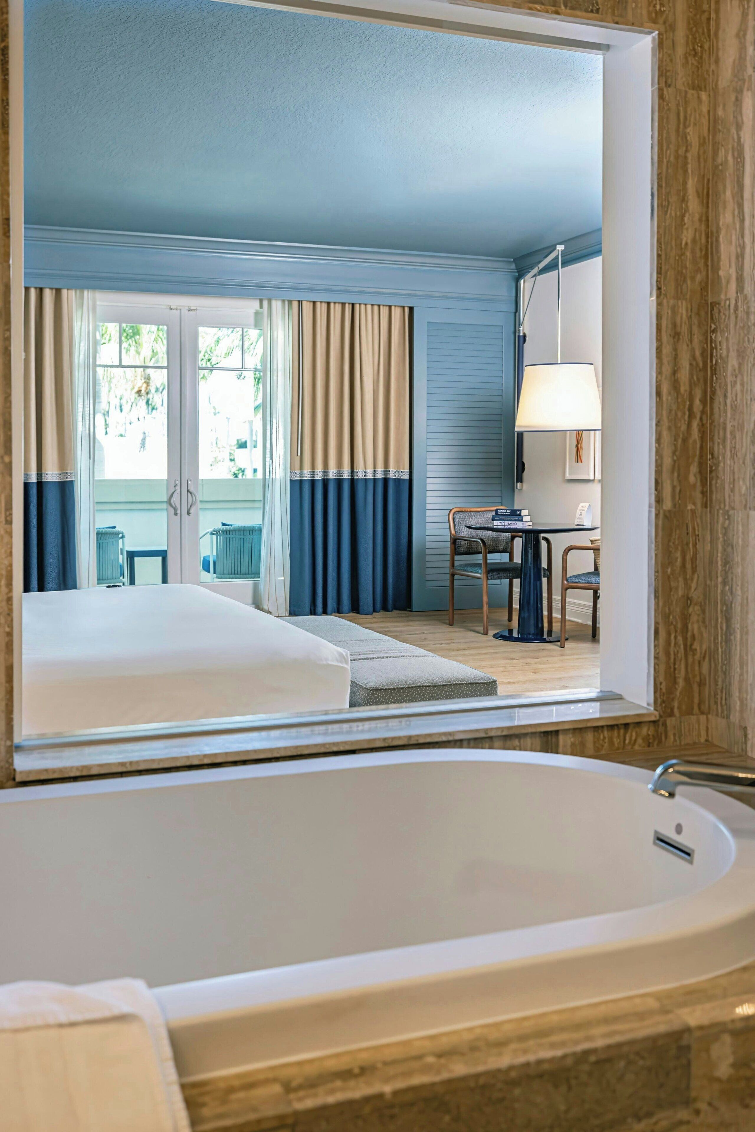 A hotel room with a large bathtub, a dining area with a table and chairs, and a view through a window with curtains.