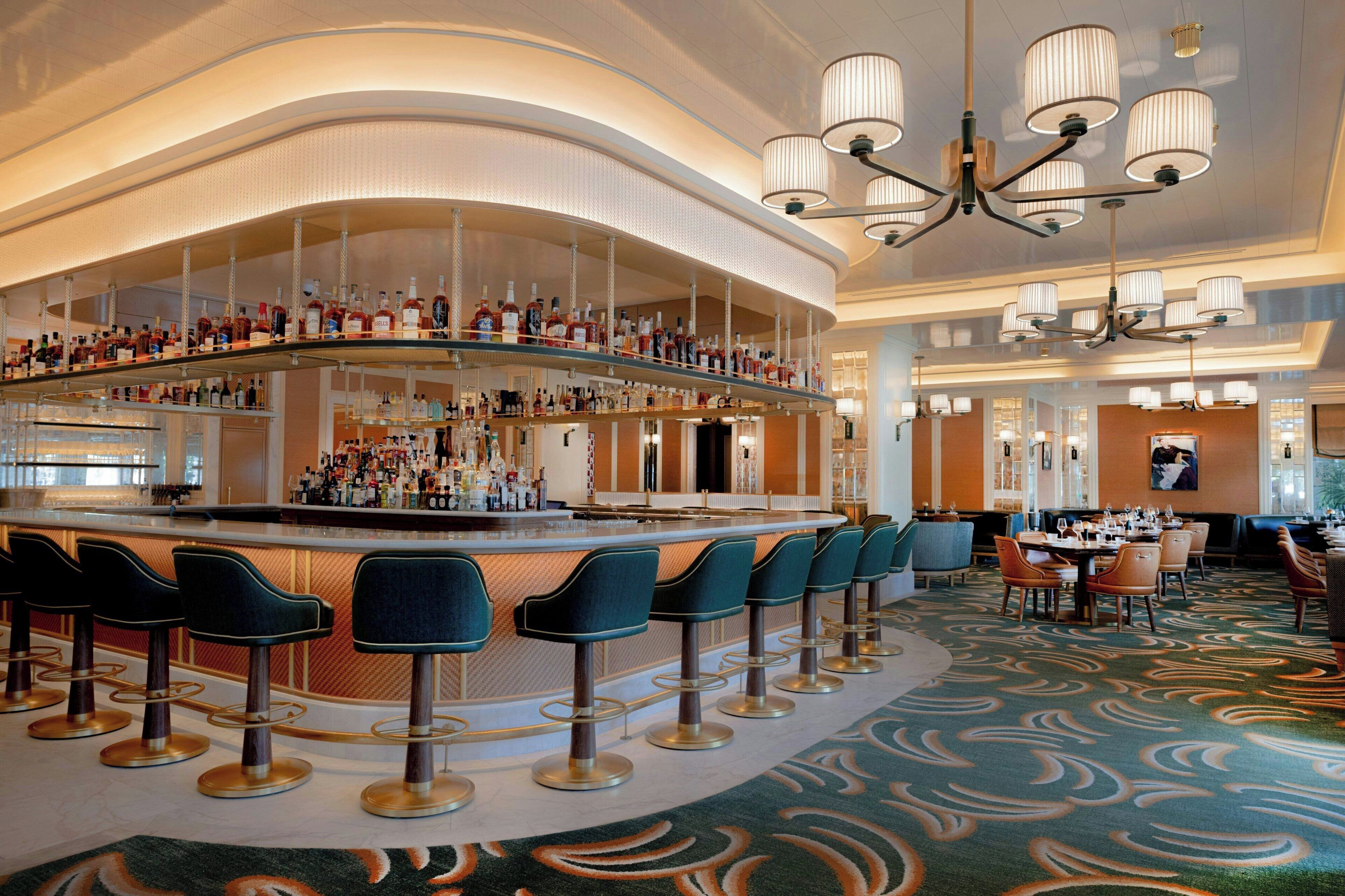 Eine Bar mit mehreren Hockern und einer gebogenen Bar, umgeben von einem gemusterten Teppich und Kronleuchtern.