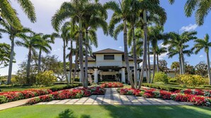A large, two-story building with a symmetrical facade, surrounded by palm trees and a well-maintained garden with red flowers and green grass. - The Seagate Hotel, Golf & Beach Club, Autograph Collection (Delray Beach)