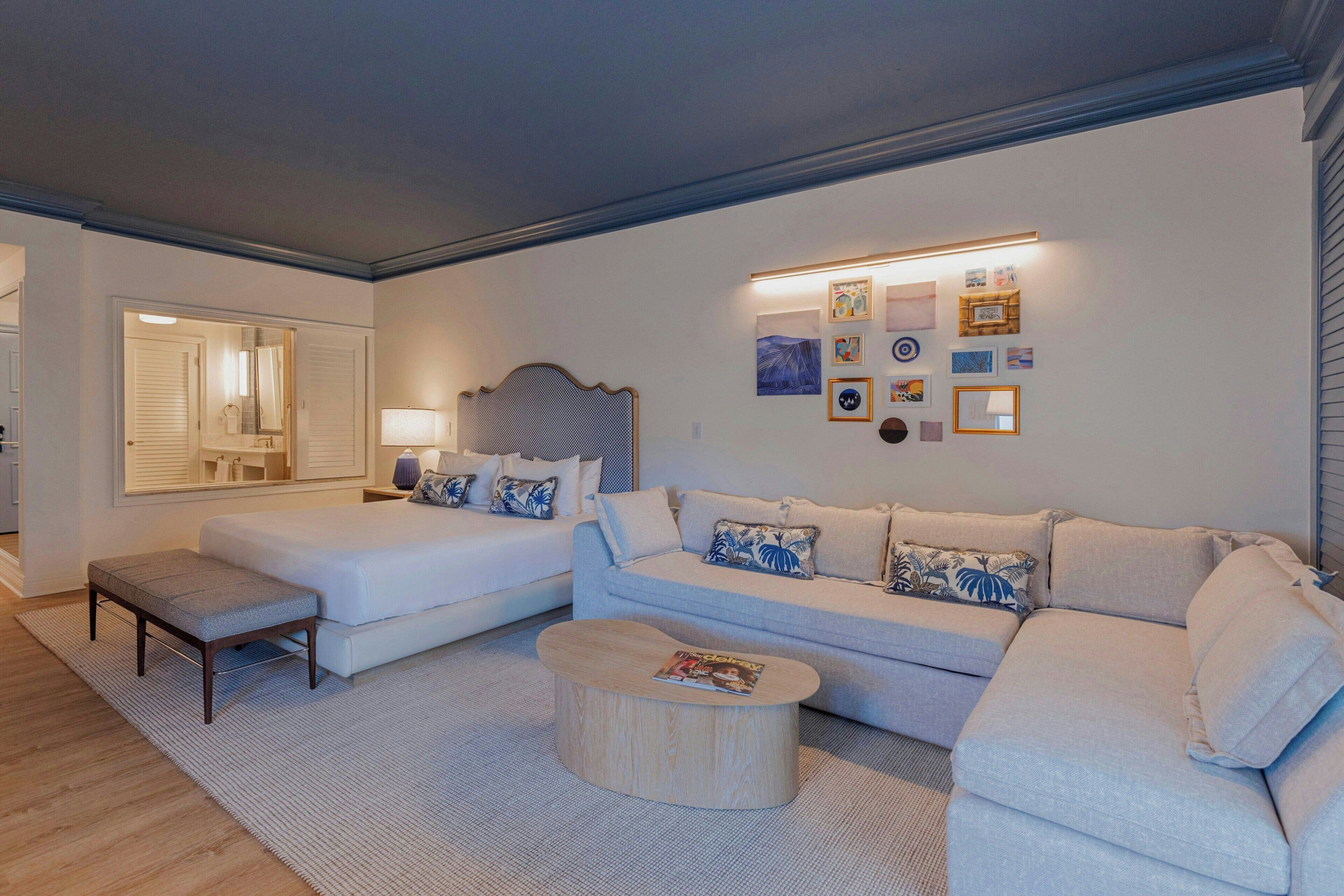 Een moderne woonkamer met een bank, een zitbankje en een salontafel. In de achtergrond staat een bed met hoofdeinde en een spiegel.