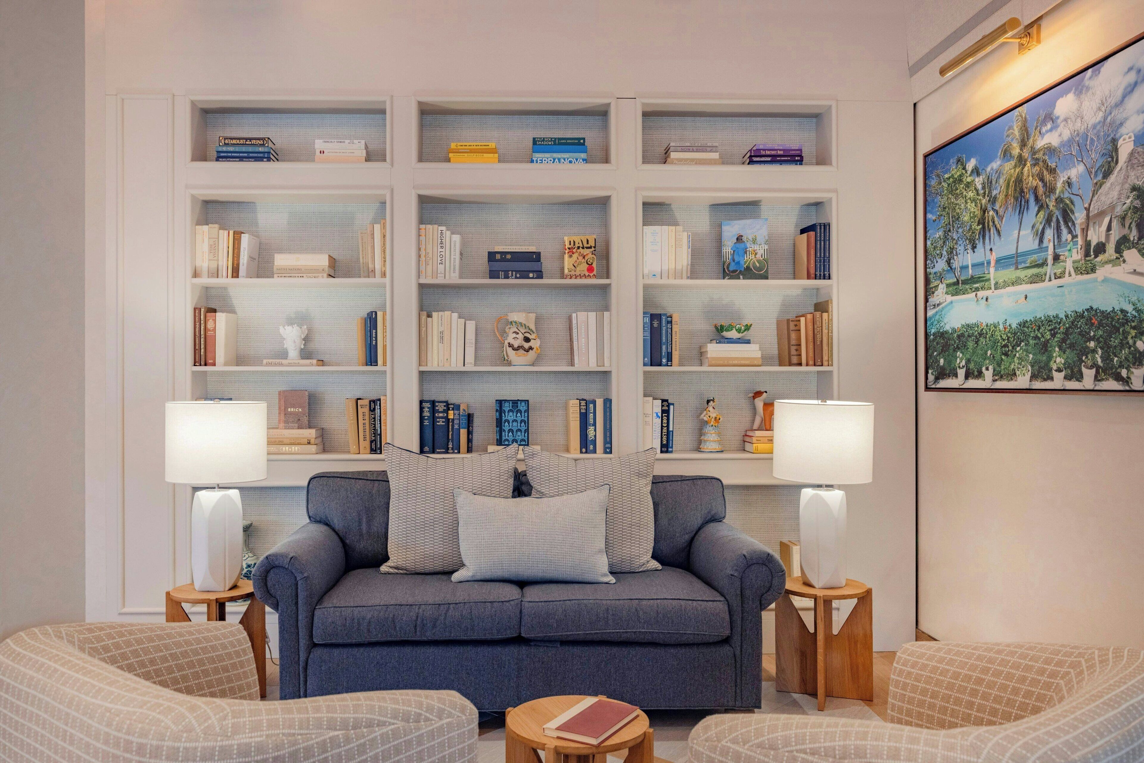 A living room with a grey sofa, two armchairs, a bookshelf filled with books, and a painting of a tropical scene.