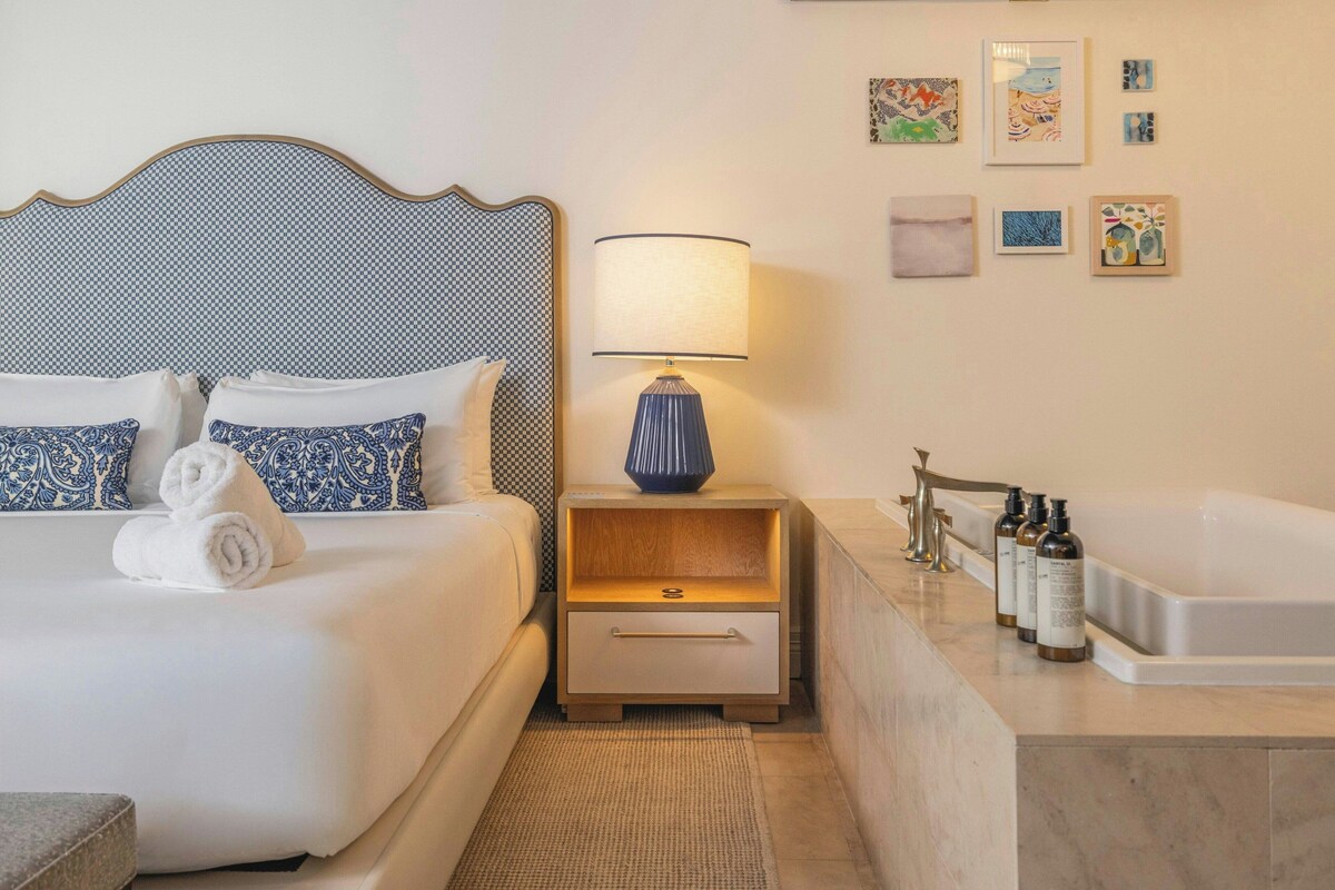 A hotel room with a bed, a nightstand with a lamp, a bathroom with a bathtub, and artwork on the wall.