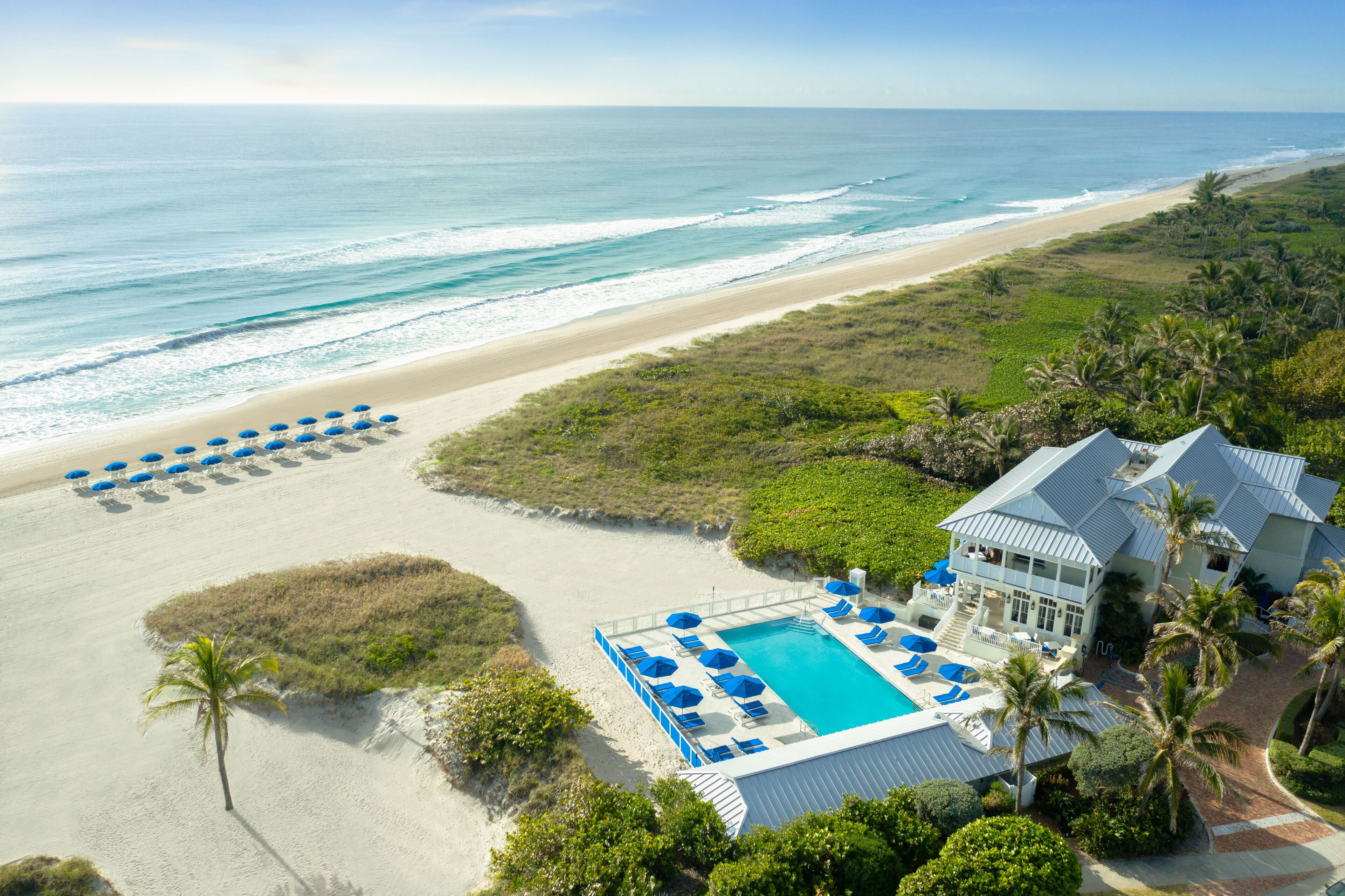 Vista aérea de un resort frente a la playa con piscina, sombrillas y una casa grande con vista al océano.