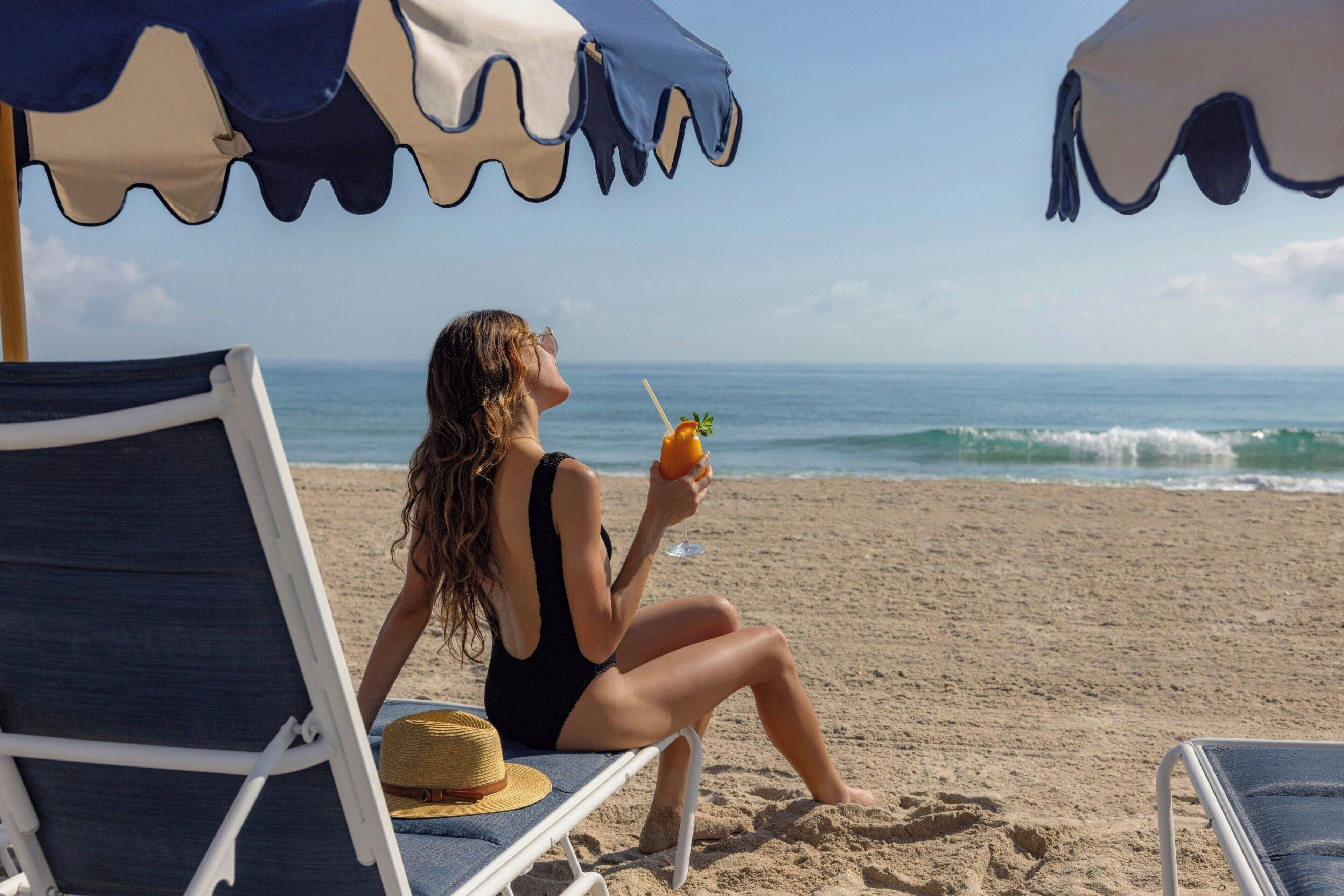 A person sitting on a beach chair under a beach umbrella, holding a drink.