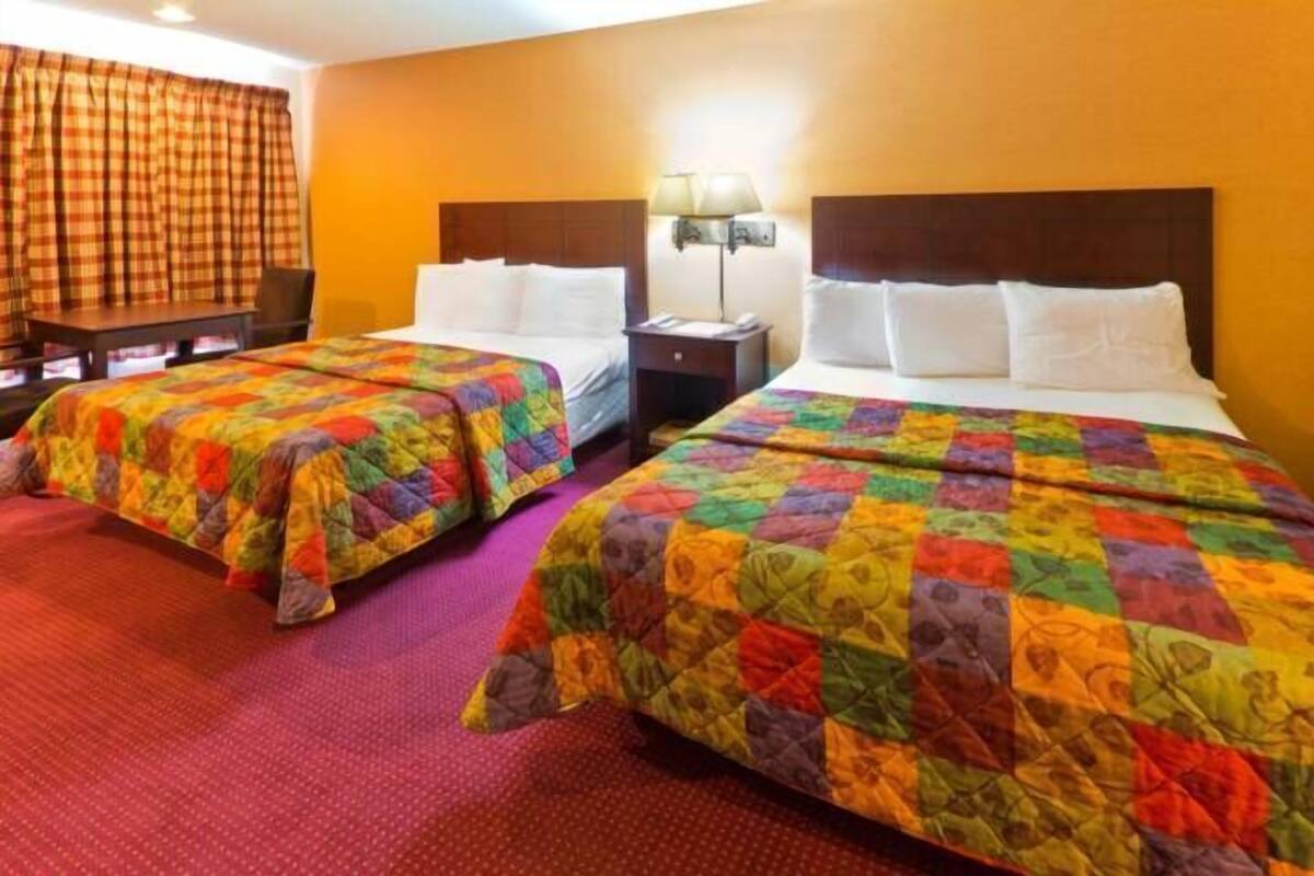 Hotellihuone, jossa on kaksi sänkyä, ja jokaisessa sängyssä on värikäs peitto. Lisäksi huoneessa on pieni pöytä, tuoli ja yöpöytä, jossa on lamppu.