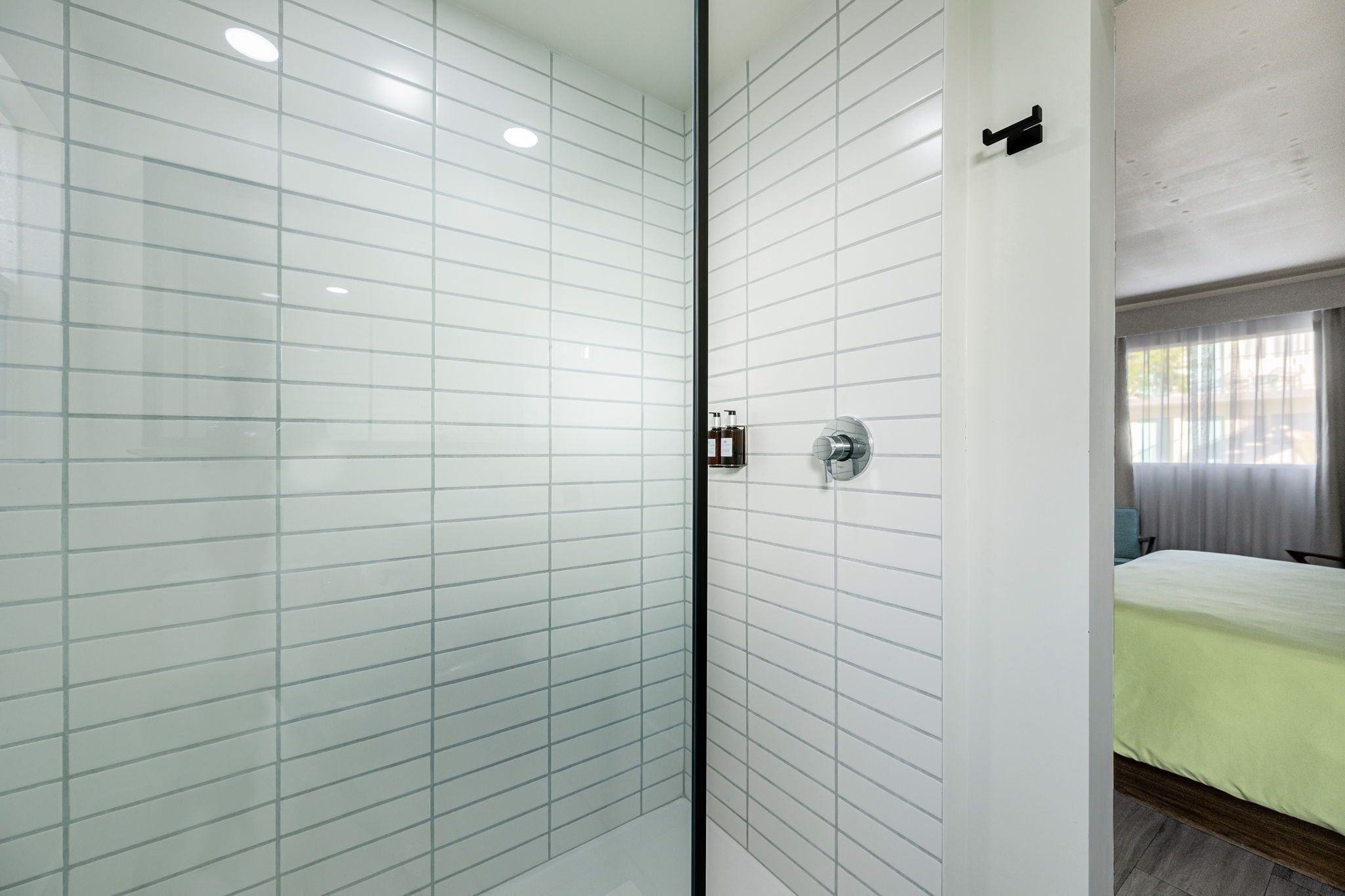 Un baño moderno con paredes de azulejos blancos, una cabina de ducha de vidrio y una cama visible con una colcha verde en la habitación contigua.