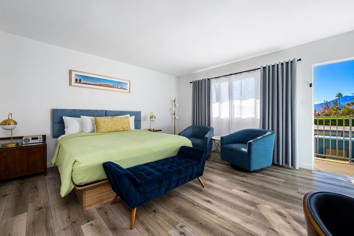 Quarto de hotel com uma cama, um banco, duas cadeiras, uma mesa de cabeceira, um rádio e um quadro na parede.
