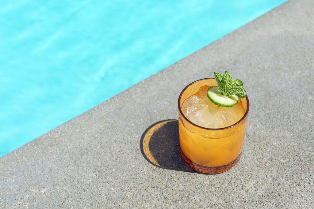 A glass of iced drink with a mint garnish on a concrete surface near a pool.