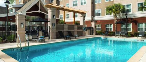 A swimming pool with lounge chairs and umbrellas, surrounded by a building with multiple windows and a covered area.