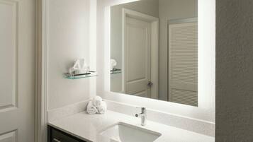 Un baño con un lavabo blanco, un estante de vidrio con toallas y un espejo que refleja una puerta y una ventana.