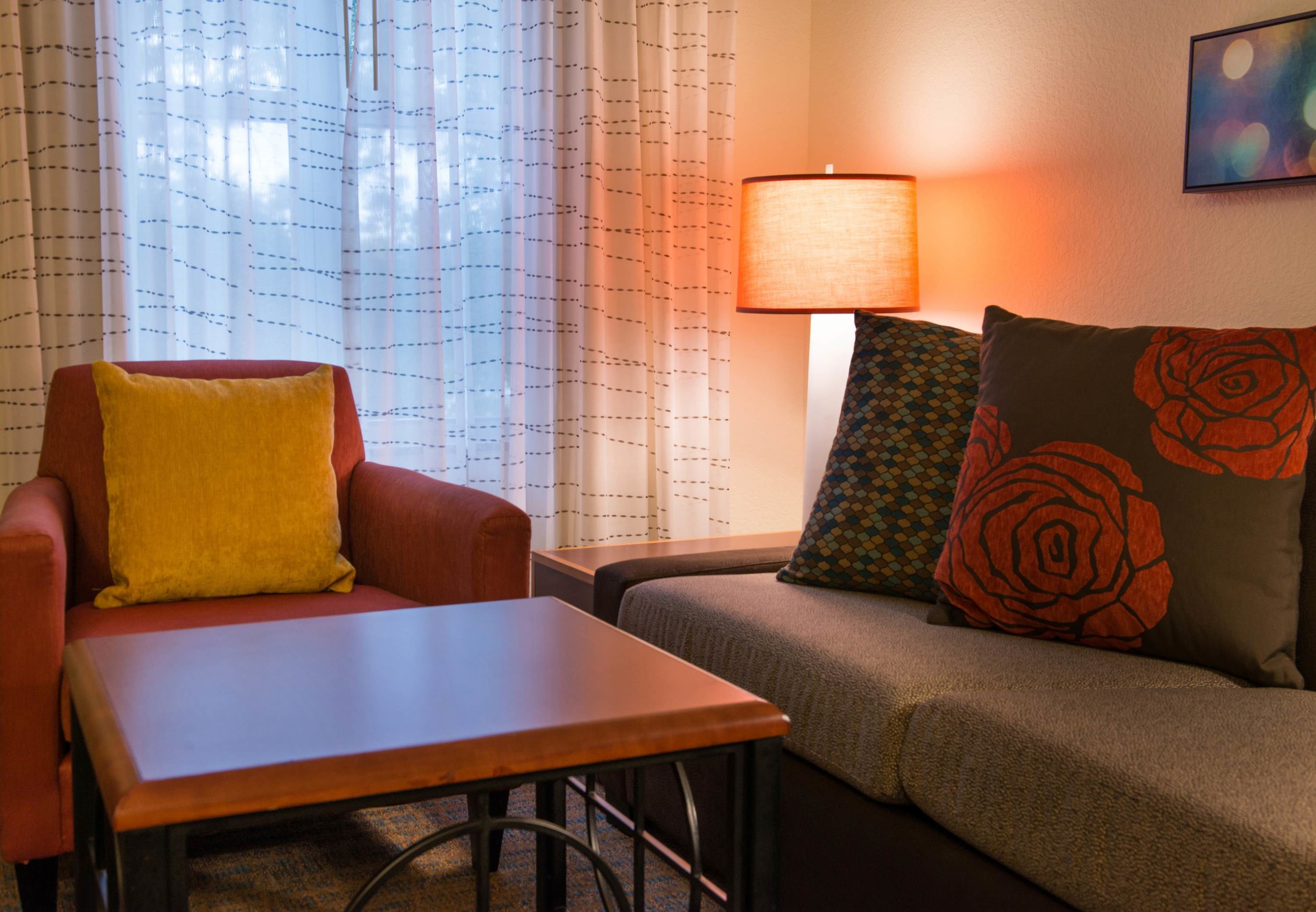 A cozy room with a red armchair, a wooden table, a sofa with patterned cushions, a lamp, and a window with sheer curtains.