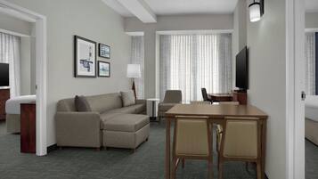 A hotel room with a sofa, a dining table with chairs, a TV, and framed pictures on the wall.