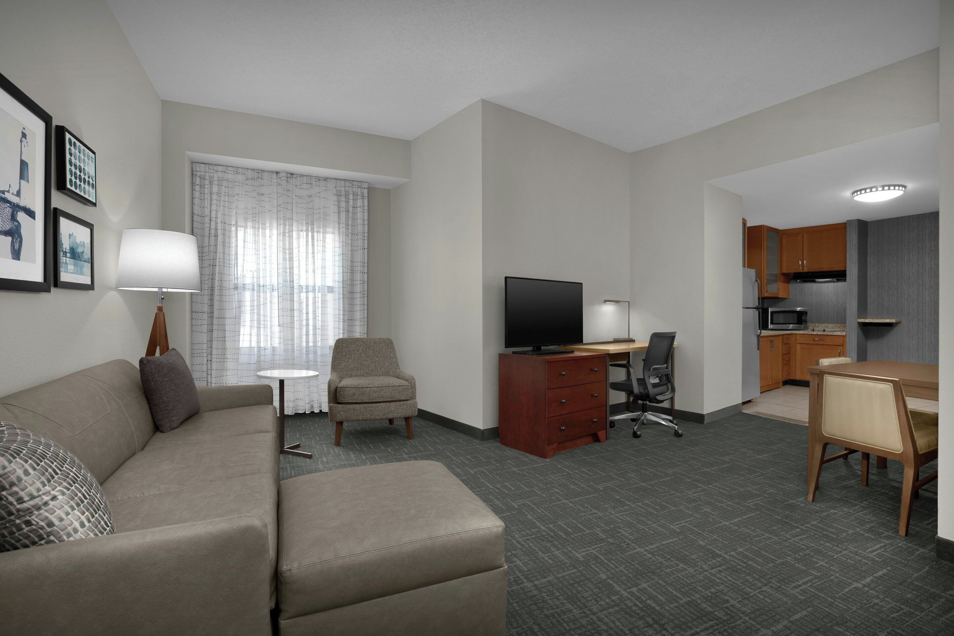 A hotel room with a sofa, a TV, a desk, a chair, a lamp, and a kitchen area.