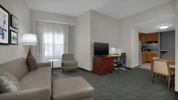 A hotel room with a sofa, a TV, a desk, a chair, a lamp, and a kitchen area.