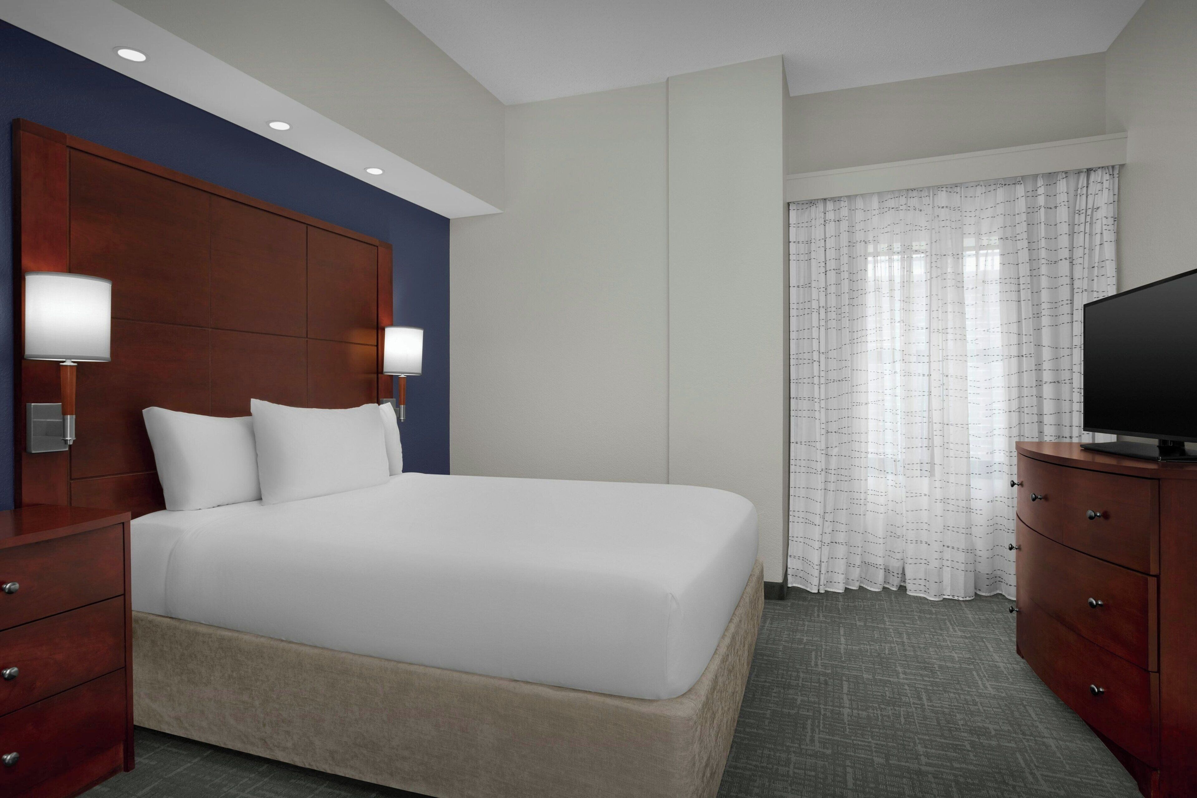 Una habitación de hotel con una cama grande, lámparas de noche, un televisor, un aparador de madera y una ventana con cortinas translúcidas.