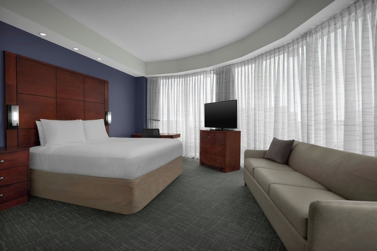 A hotel room with a large bed, a sofa, a TV on a wooden cabinet, and a window with sheer curtains.