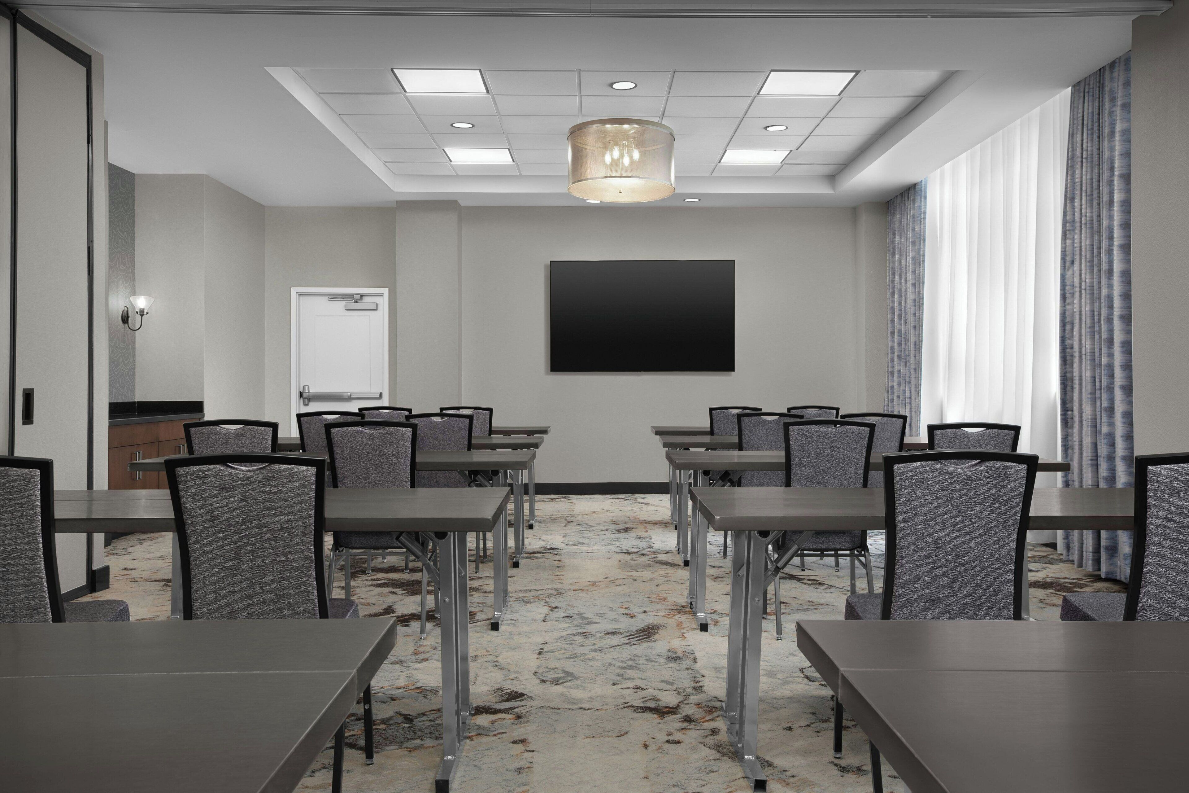 Un salón de conferencias con una gran pantalla plana, varias mesas y sillas, una alfombra estampada e iluminación empotrada en el techo.