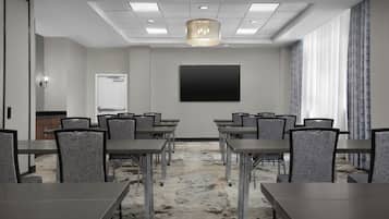 Un salón de conferencias con una gran pantalla plana, varias mesas y sillas, una alfombra estampada e iluminación empotrada en el techo.