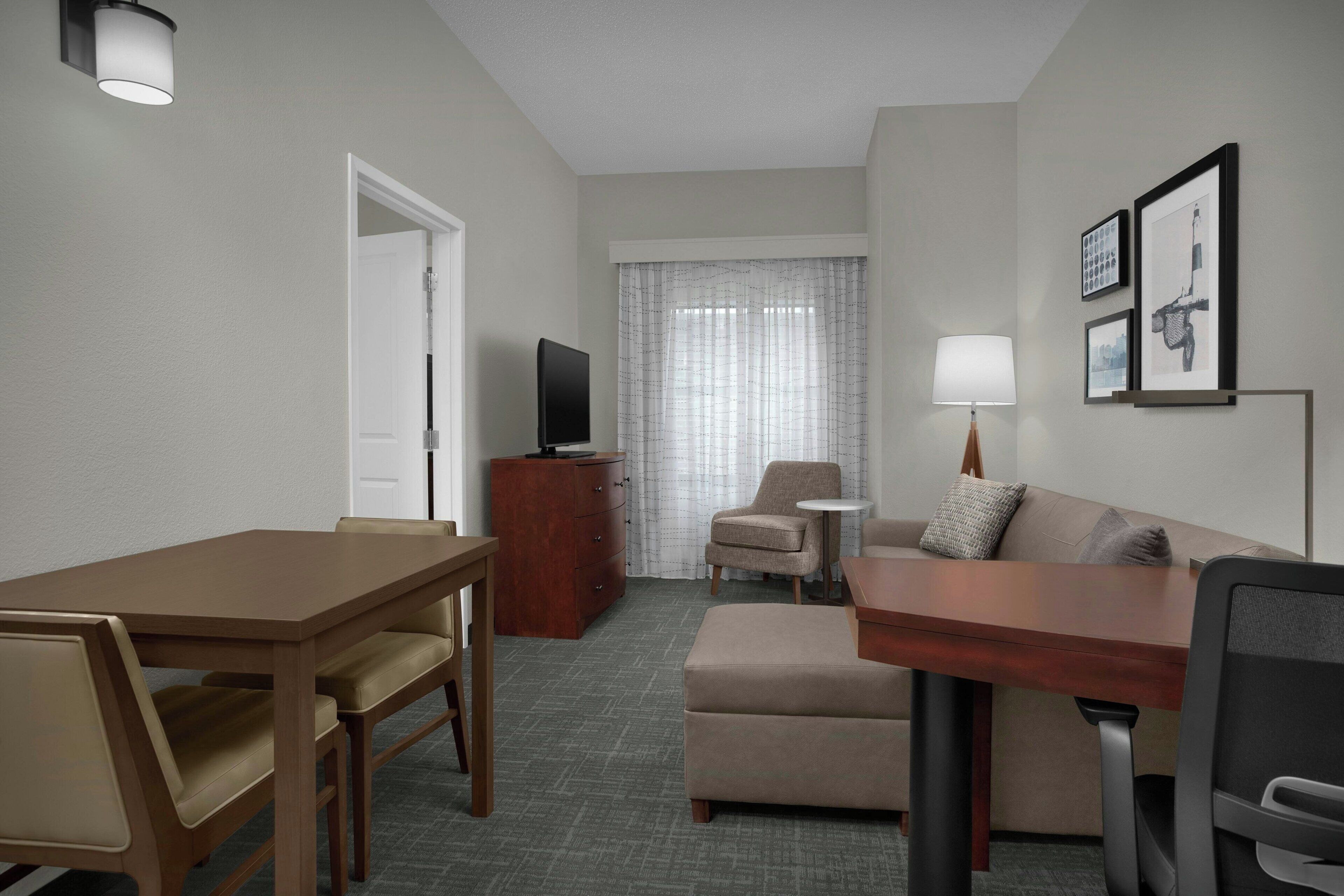 A hotel room with a wooden dining table, a sofa, a TV, a small side table, and a lamp.