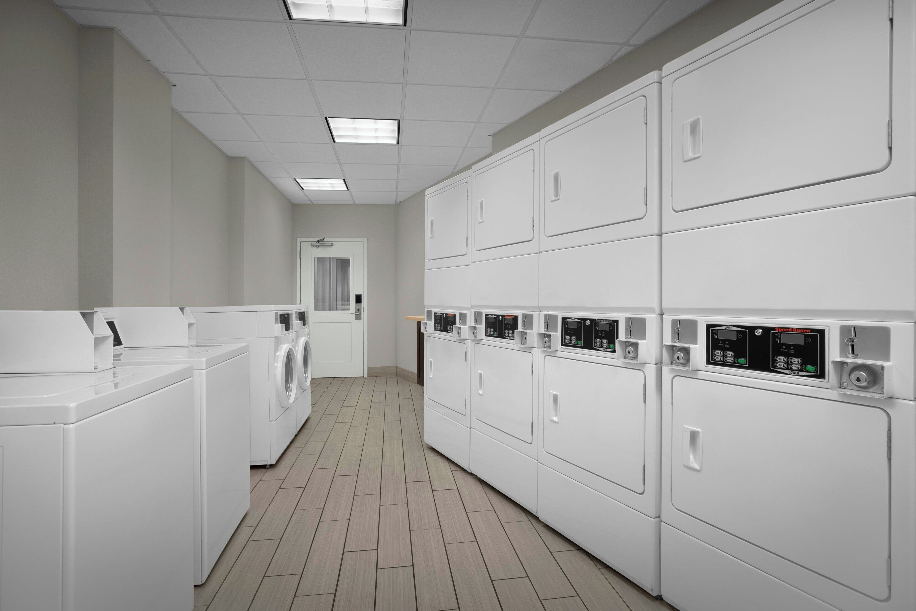 A laundry facility with multiple washing machines and dryers, a tiled floor, and a door at the end of the room.
