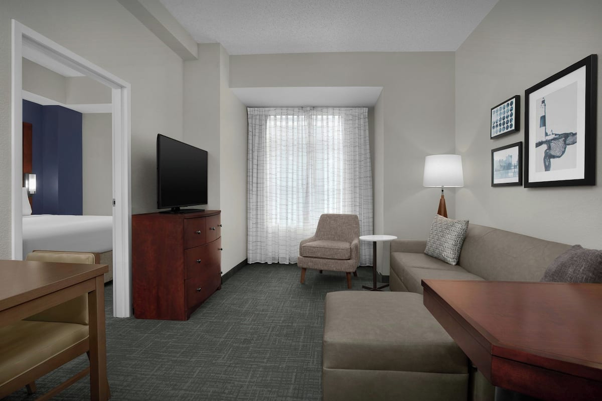 A hotel room with a bed, a TV, a desk, a chair, a sofa, a small table, and a window with curtains.