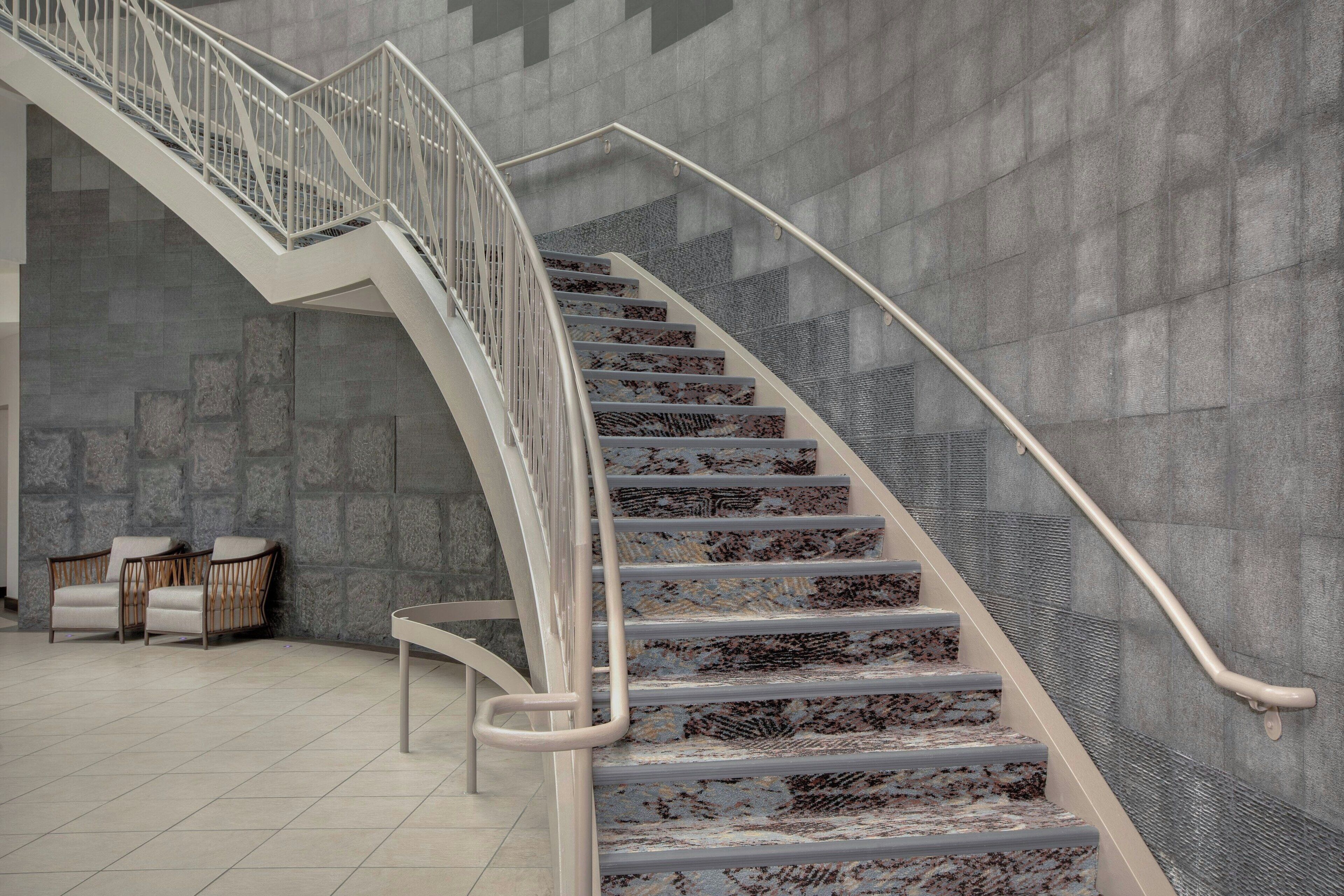 Una escalera moderna con barandilla de metal, paredes de piedra y un rincón de descanso con sillas.