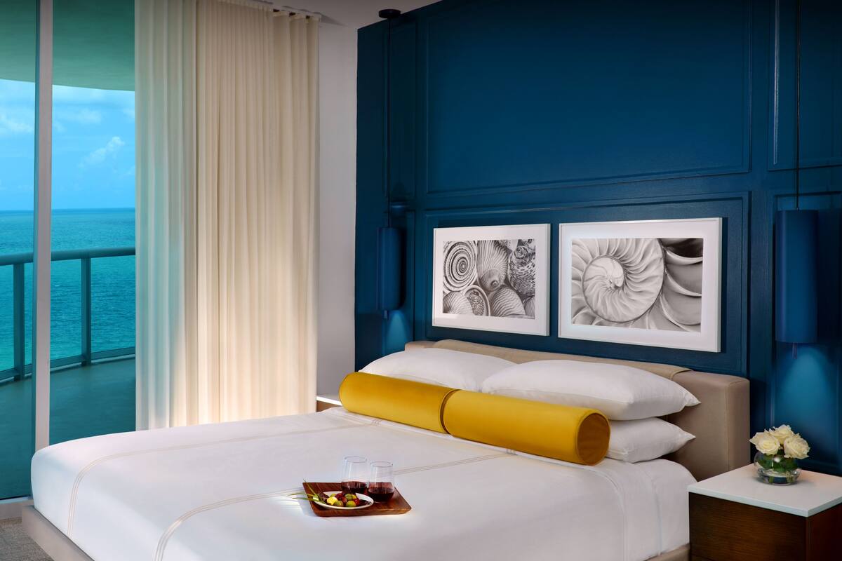 A hotel room with a large window overlooking the sea, a bed with white linens, a yellow pillow, and two framed artworks on the wall.