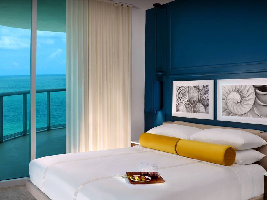 Habitación de hotel con un ventanal grande con vistas al mar, una cama con ropa blanca, una almohada amarilla y dos obras de arte enmarcadas en la pared.
