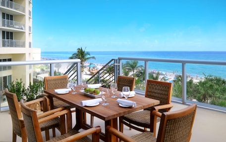 A wooden table with four chairs, a plate with two apples, and several wine glasses on a balcony overlooking a beach with palm trees and a clear blue sky.
