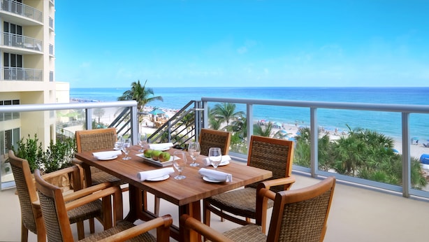 Een houten tafel met vier stoelen, een bord met twee appels en verschillende wijnglazen op een balkon met uitzicht op een strand met palmbomen en een heldere blauwe lucht.