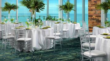 A banquet hall with round tables, clear chairs, and white tablecloths.