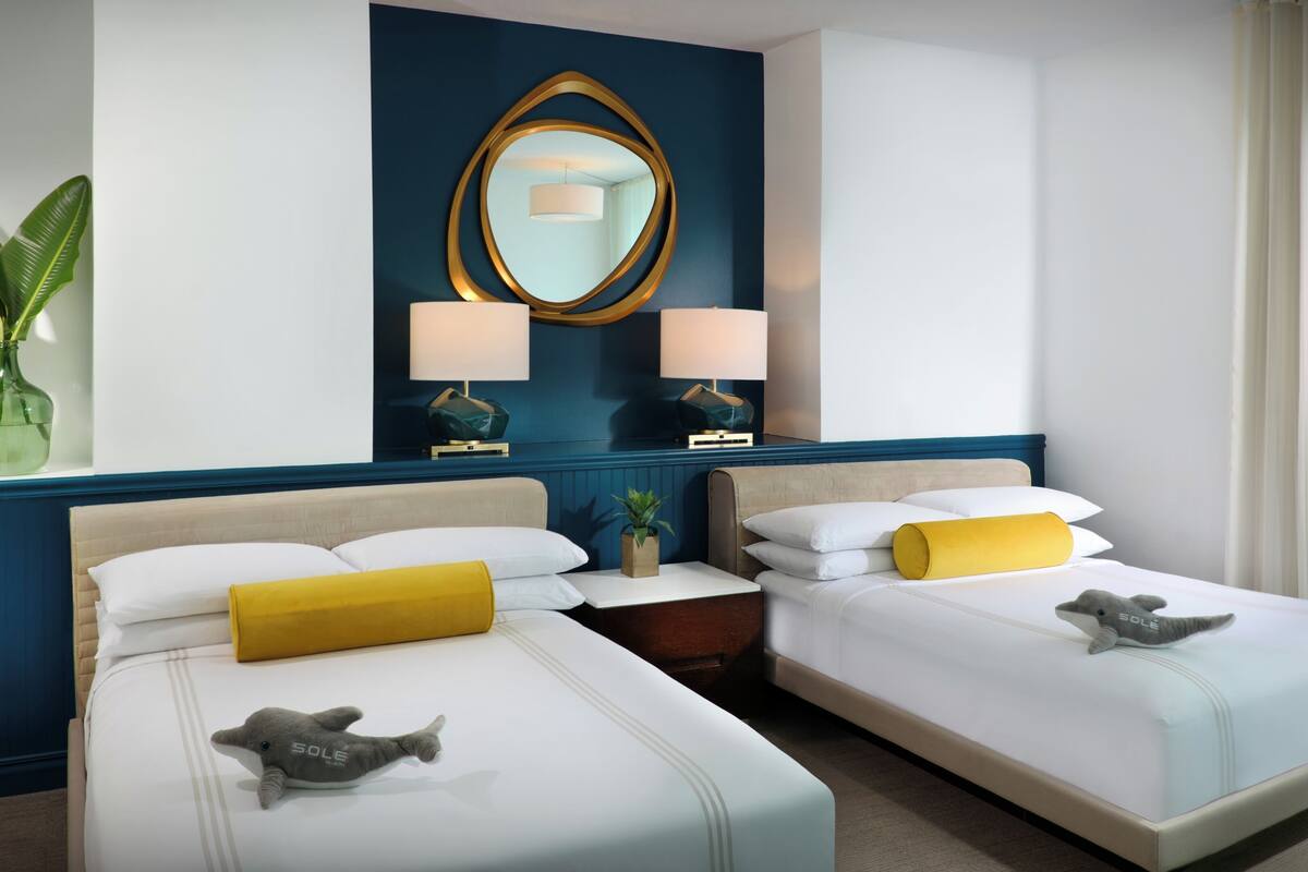 A hotel room with two beds, a large mirror, bedside lamps, a small plant, and a window with curtains.