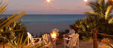 A beachside seating area with two Adirondack chairs, a small fire pit, and a string of lights.