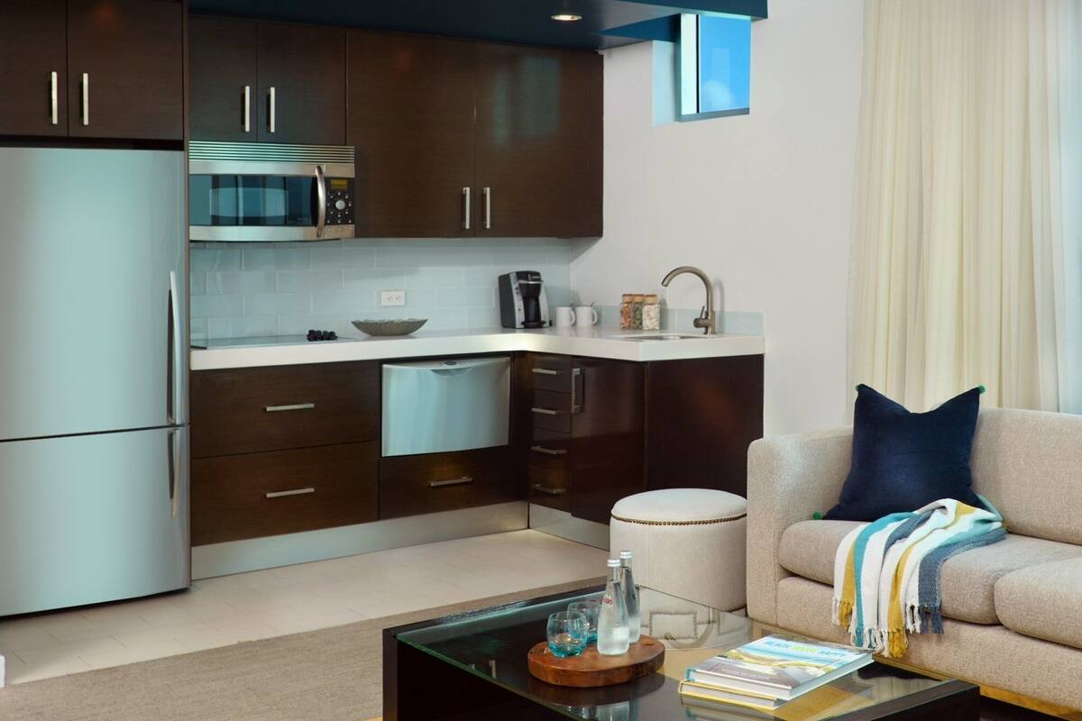 A modern kitchen with dark cabinets, a stainless steel refrigerator, and a microwave. There is a dining area with a glass coffee table and a sofa with a throw blanket.