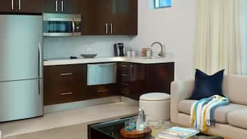 A modern kitchen with dark cabinets, a stainless steel refrigerator, and a microwave. There is a dining area with a glass coffee table and a sofa with a throw blanket.
