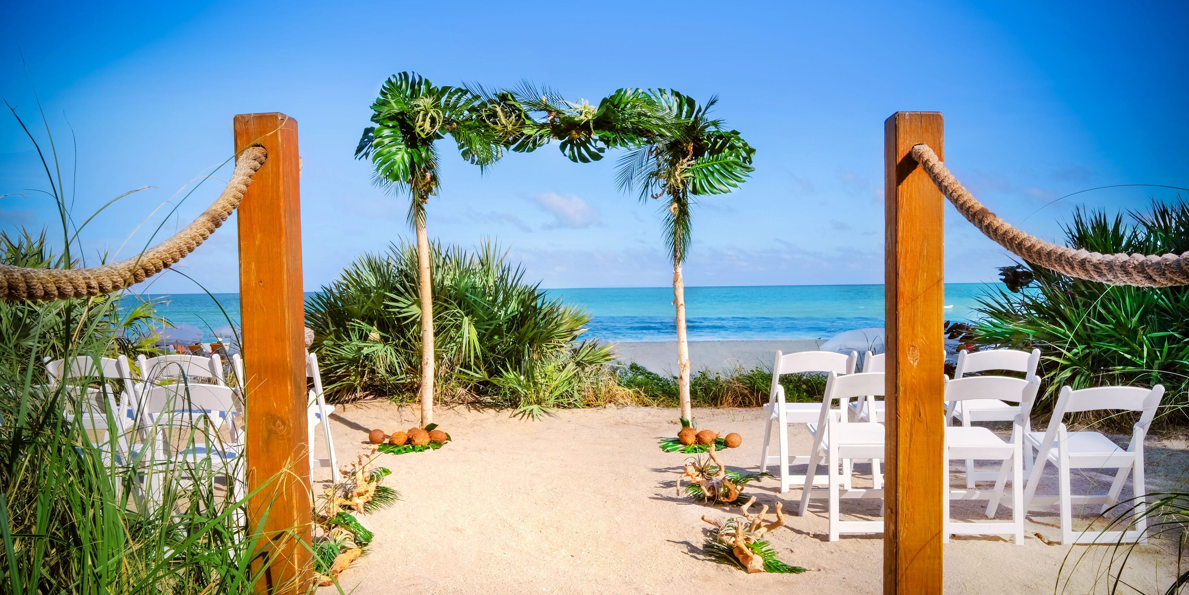 A beach wedding setup with a wooden archway, white chairs, and palm trees.