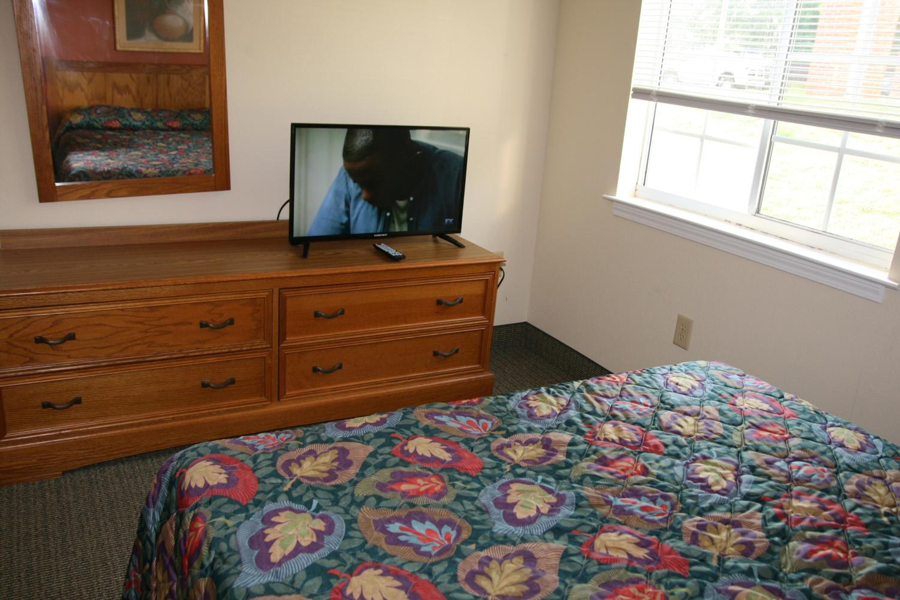 A bedroom with a wooden dresser, a television on top, a bed with a floral bedspread, and a window with blinds.