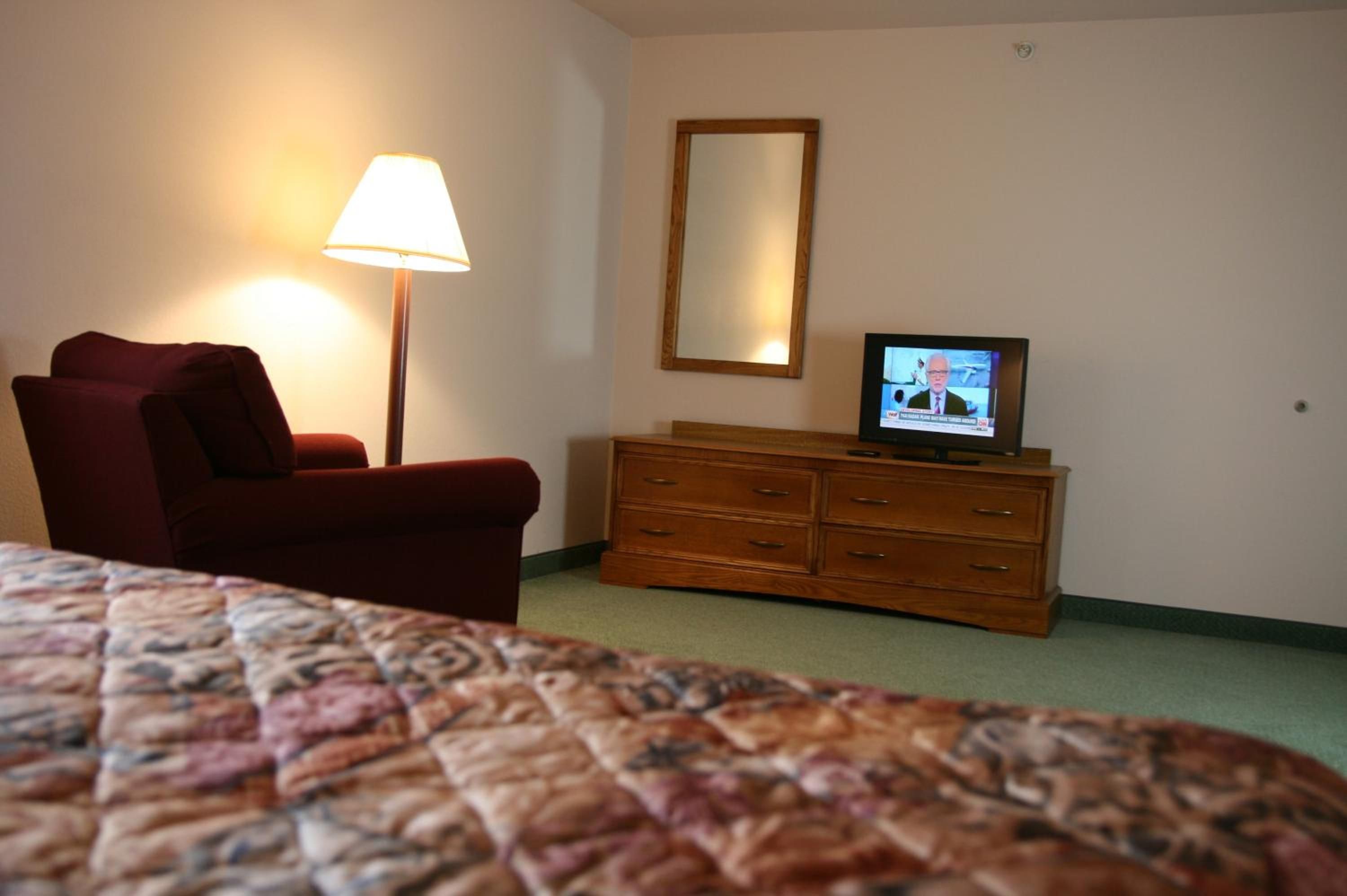 A hotel room with a bed, a red armchair, a wooden dresser, a television, and a floor lamp.