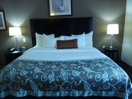 Una cama bien hecha con una colcha estampada, dos lámparas de noche, una mesita de noche con un teléfono y un reloj, y un cuadro en la pared.