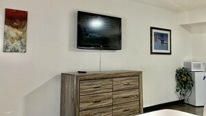 Quarto com televisão de tela plana na parede, aparador de madeira com gavetas, quadro emoldurado e geladeira branca.