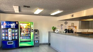 Uma área de recepção limpa e moderna, com máquina de refrigerantes Pepsi, caixa eletrônico e uma pequena planta no balcão.