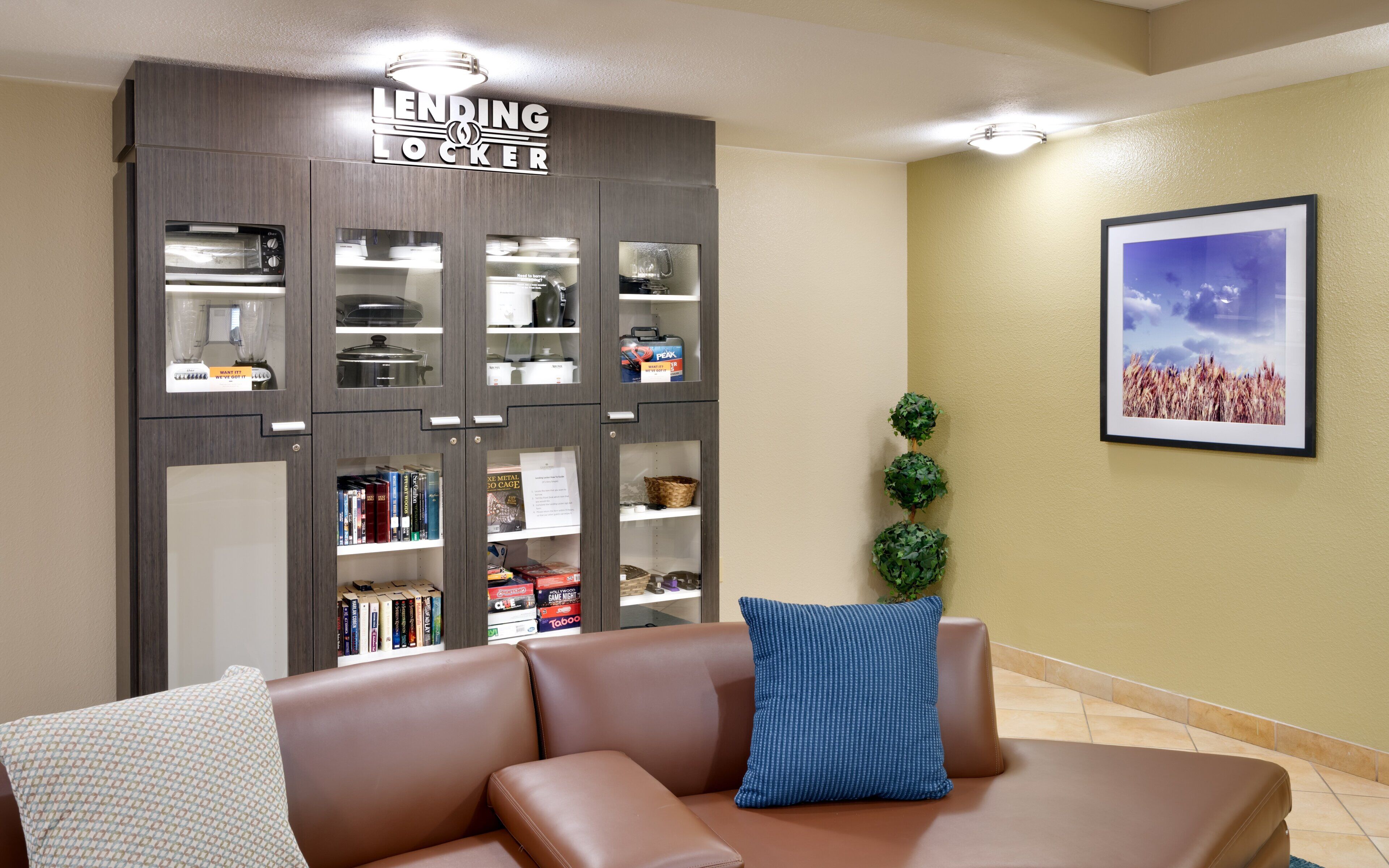 Una habitación con un sofá de cuero marrón, un armario 'Lending Locker' con varios objetos, un cuadro en la pared y una planta pequeña.