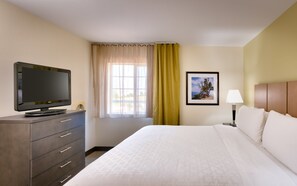 A hotel room with a bed, a television on a dresser, a window with curtains, a framed picture on the wall, and a bedside lamp. - Candlewood Suites Dallas Plano East Richardson by IHG (Plano)