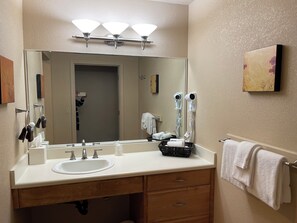 A bathroom with a large mirror, three wall-mounted lights, a sink, two wooden drawers, a towel rack with a white towel, and a hairdryer on the wall.