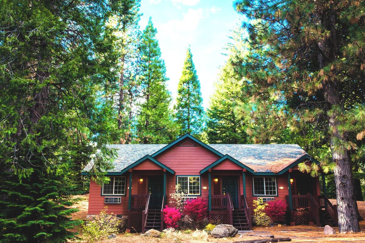A red cabin with a steep roof, surrounded by tall trees and a clear sky.