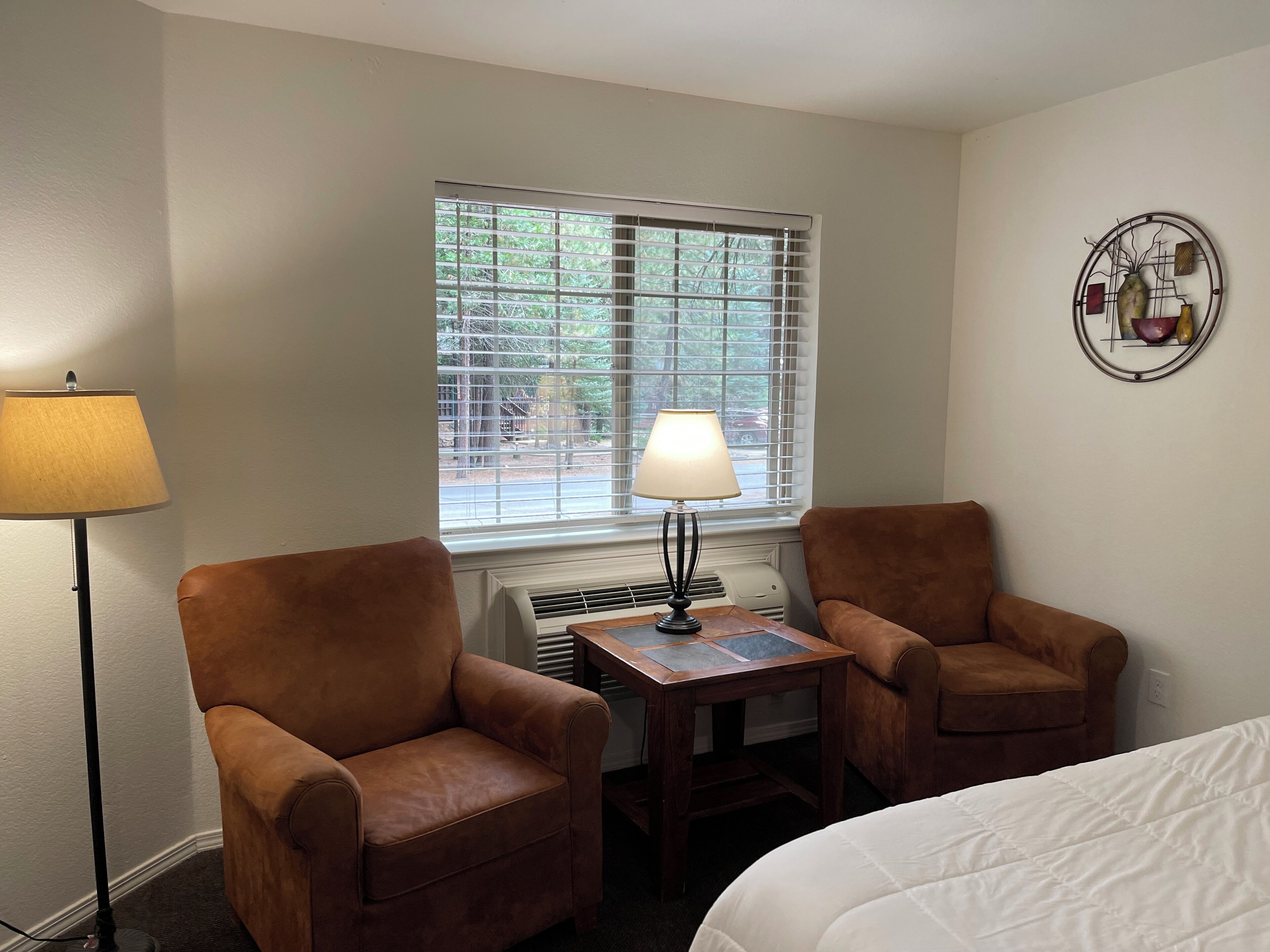 A room with two brown leather armchairs, a small wooden table, a white bed, a window with blinds, and a wall-mounted decorative piece.