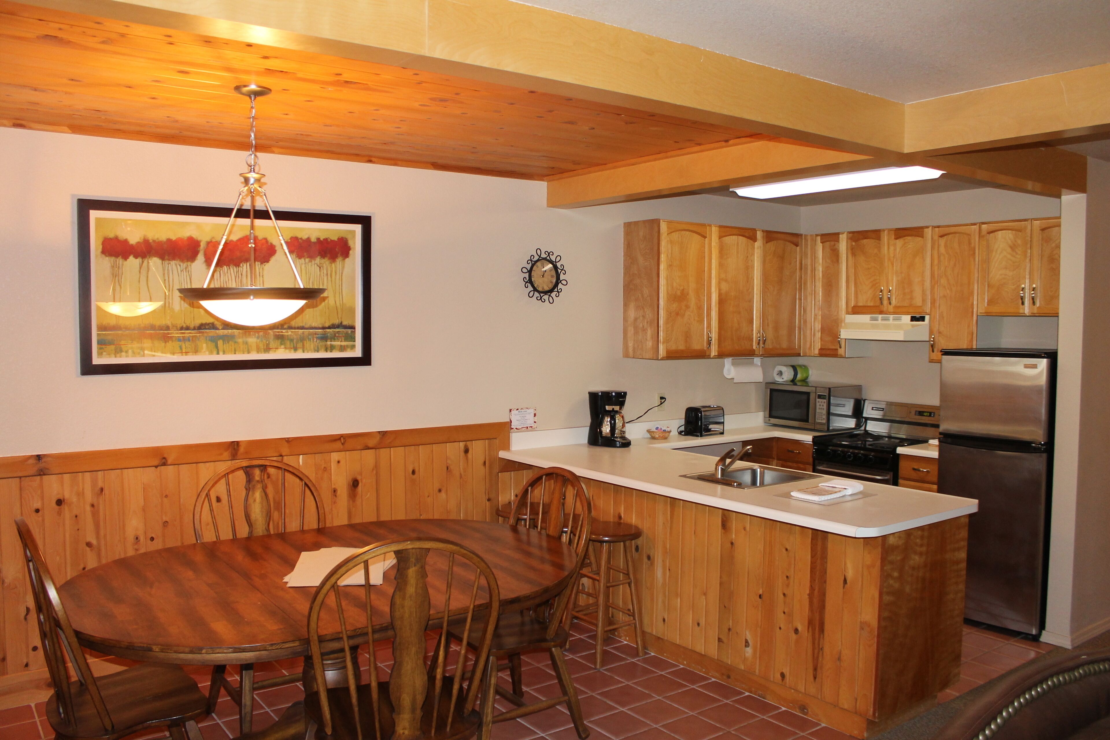 A kitchen with wooden cabinets, a dining table with chairs, a hanging light fixture, and a clock on the wall.
