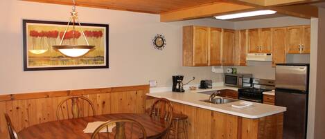 A kitchen with wooden cabinets, a dining table with chairs, a hanging light fixture, and a clock on the wall.